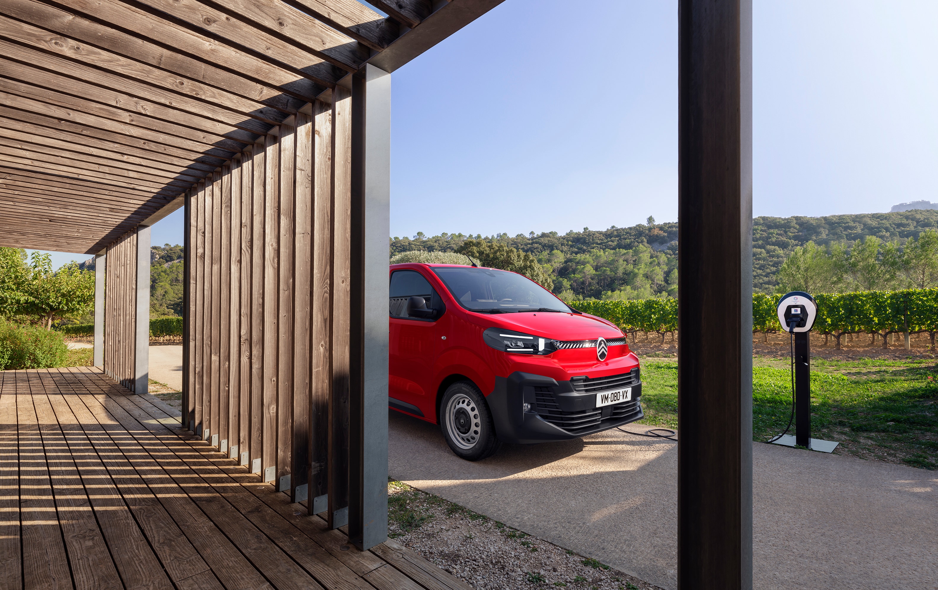 Vista exterior tres cuartos lateral de un Citroën ë-Jumpy rojo aparcado frente a un edificio de madera, mostrando diseño eléctrico moderno, practicidad diaria y presencia profesional segura.