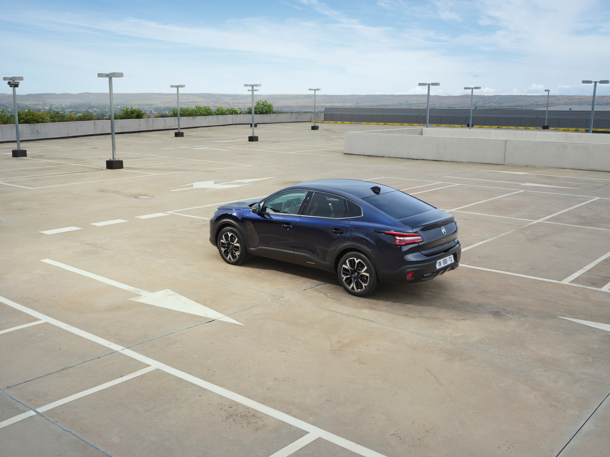 Vista trasera tres cuartos del Citroën ë-C4 X en parking de azotea abierto destacando líneas fastback elegantes para una movilidad asequible.